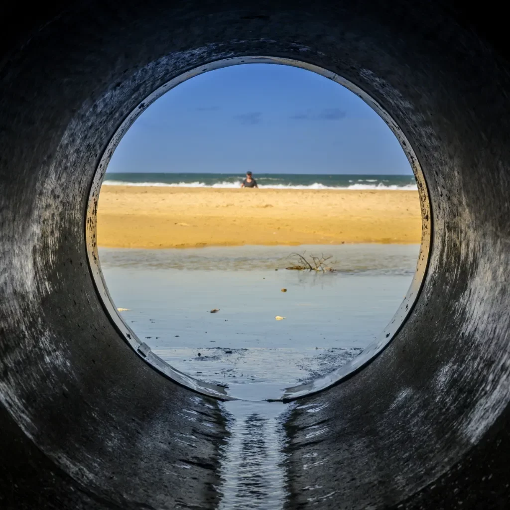 Soleco - Piaszczysta plaża i ocean widziane przez otwór dużej betonowej rury drenażowej, ilustrujące montaż systemu rur.
